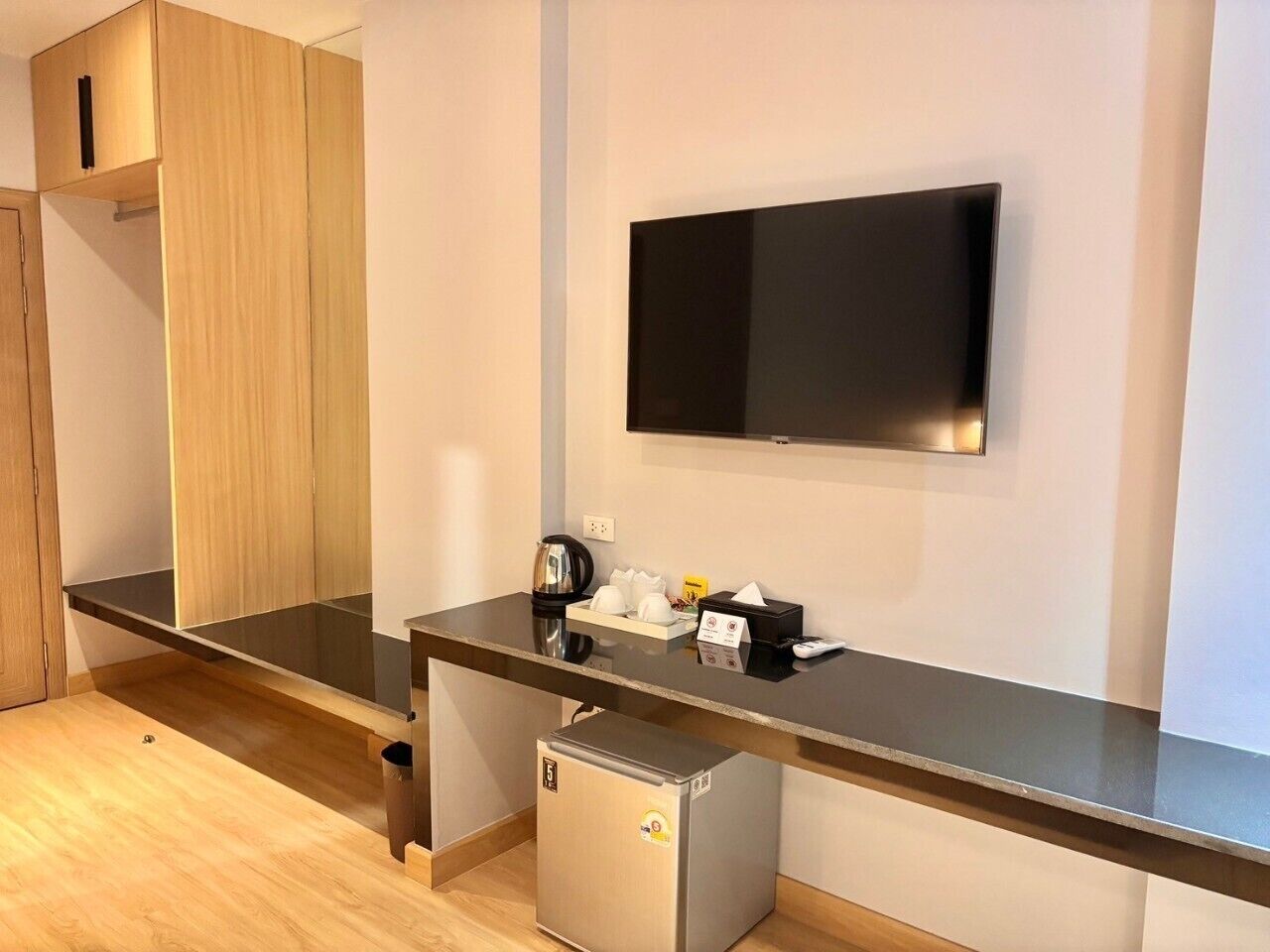 Quarto de hotel moderno com televisão de tela plana na parede, minibar, cafeteira e lixeira.