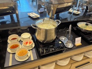 A buffet setup with a pot of boiling rice, bowls of condiments, and stacked plates. - The Essence HOTEL Chanthaburi (Chanthaburi)