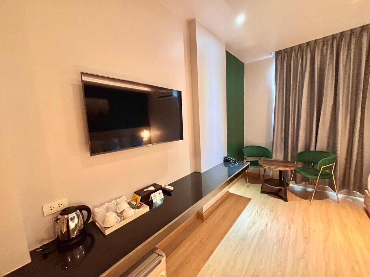 Una habitación de hotel moderna con una televisión de pantalla plana montada en la pared, una kitchenette pequeña con cafetera, una zona de comedor con mesa y sillas, y un ventanal con cortinas.