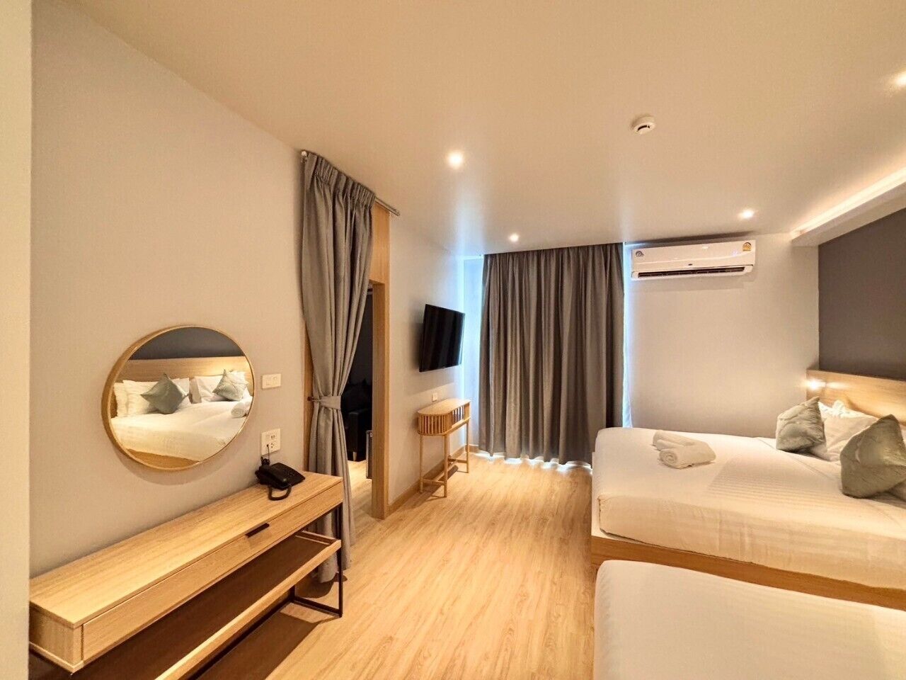 A hotel room with a bed, a bedside table, a mirror, a TV, and an air conditioning unit.