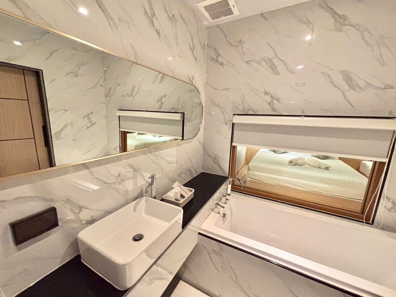 A modern bathroom with a marble vanity, a large mirror, and a window with a view of a bed.