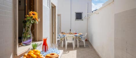 A small outdoor dining area with a table set for two, a vase with flowers, and a fruit basket on the windowsill.