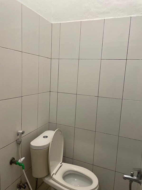 A small, clean bathroom with white tiles, a toilet, a green and white water tap, and a shower head.