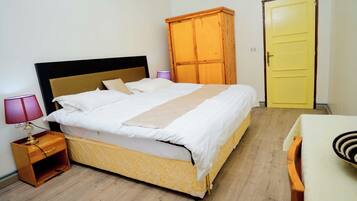 A neatly made bed with a headboard, a wooden wardrobe, and a yellow door in a simple room with wooden flooring.