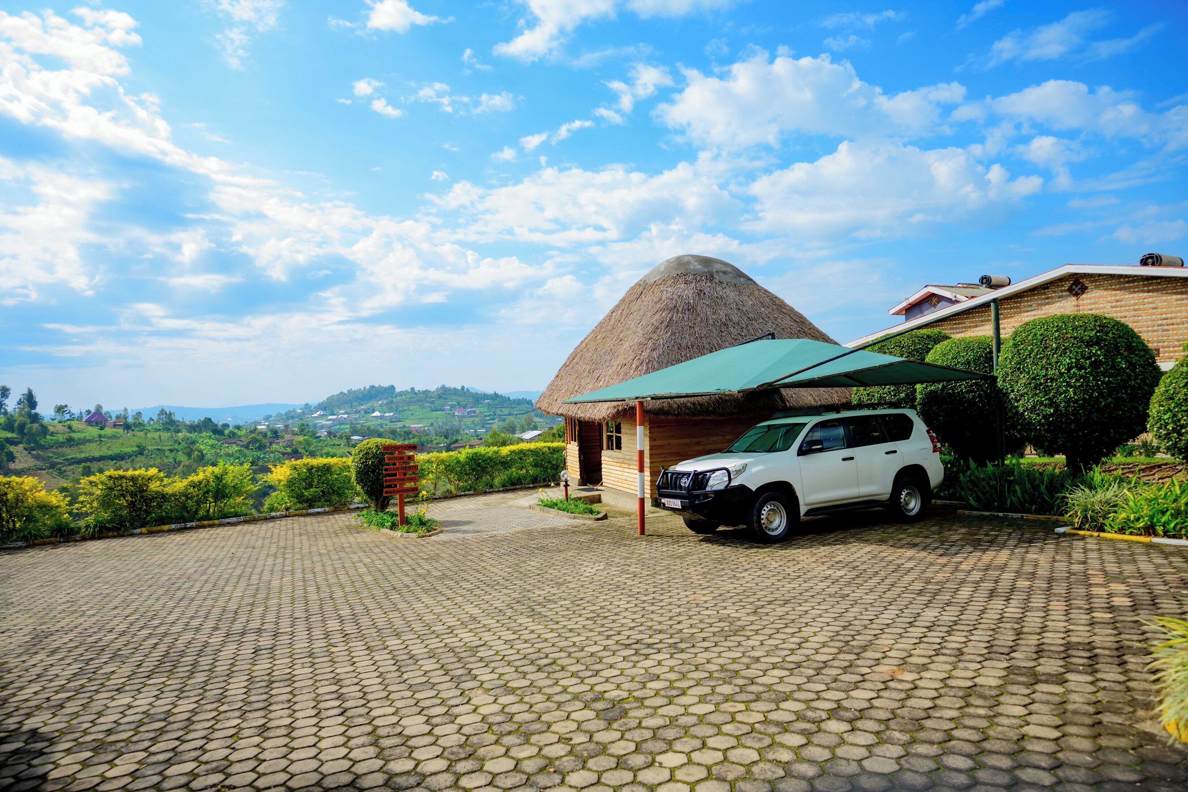 A white SUV parked in front of a thatched-roof building with a green awning, set on a cobblestone driveway overlooking a lush valley.
