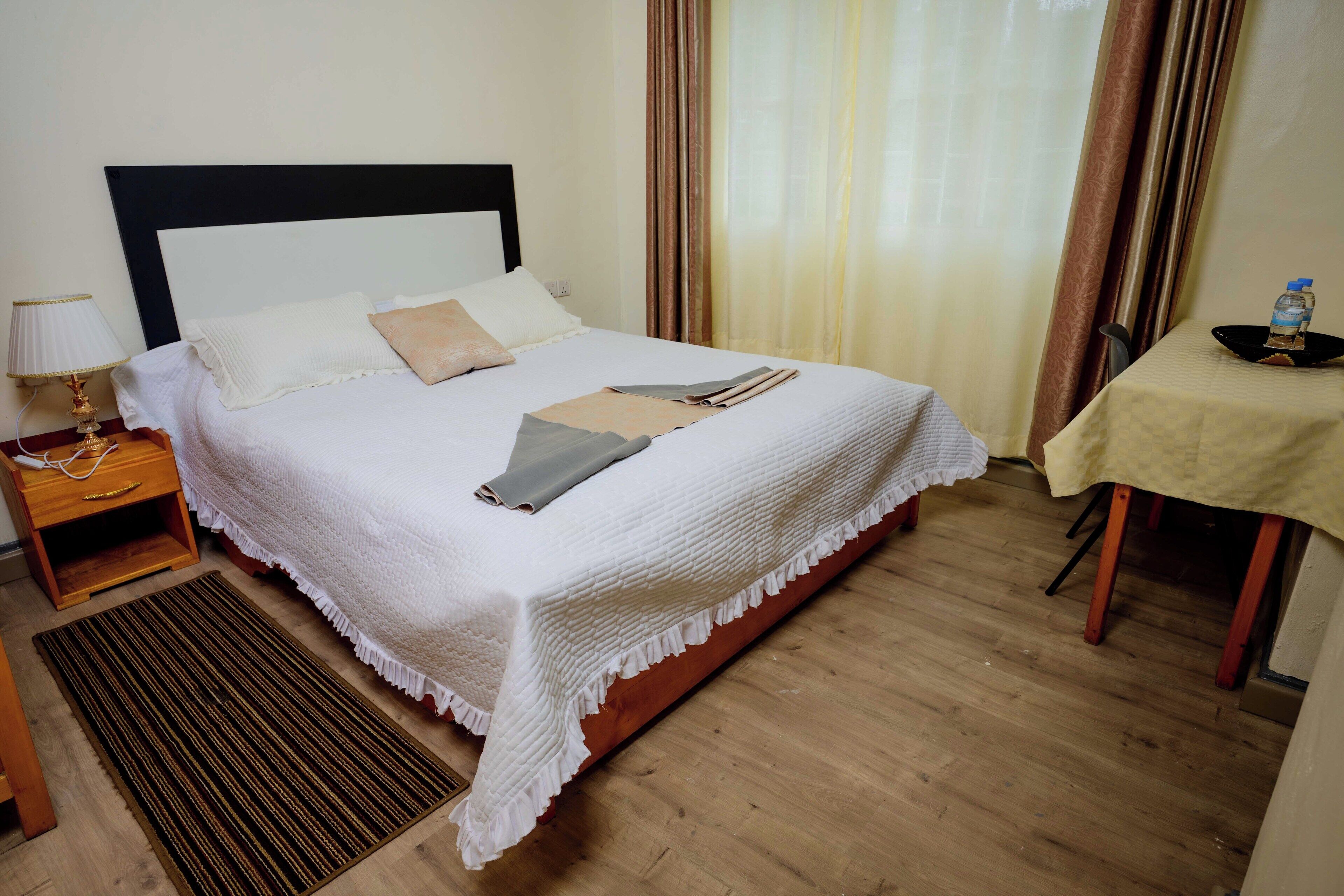 Una cama bien hecha con edredón blanco y almohadas, una mesita de noche con lámpara, una mesita auxiliar de madera, una pequeña mesa con una botella de agua, y suelo de madera.