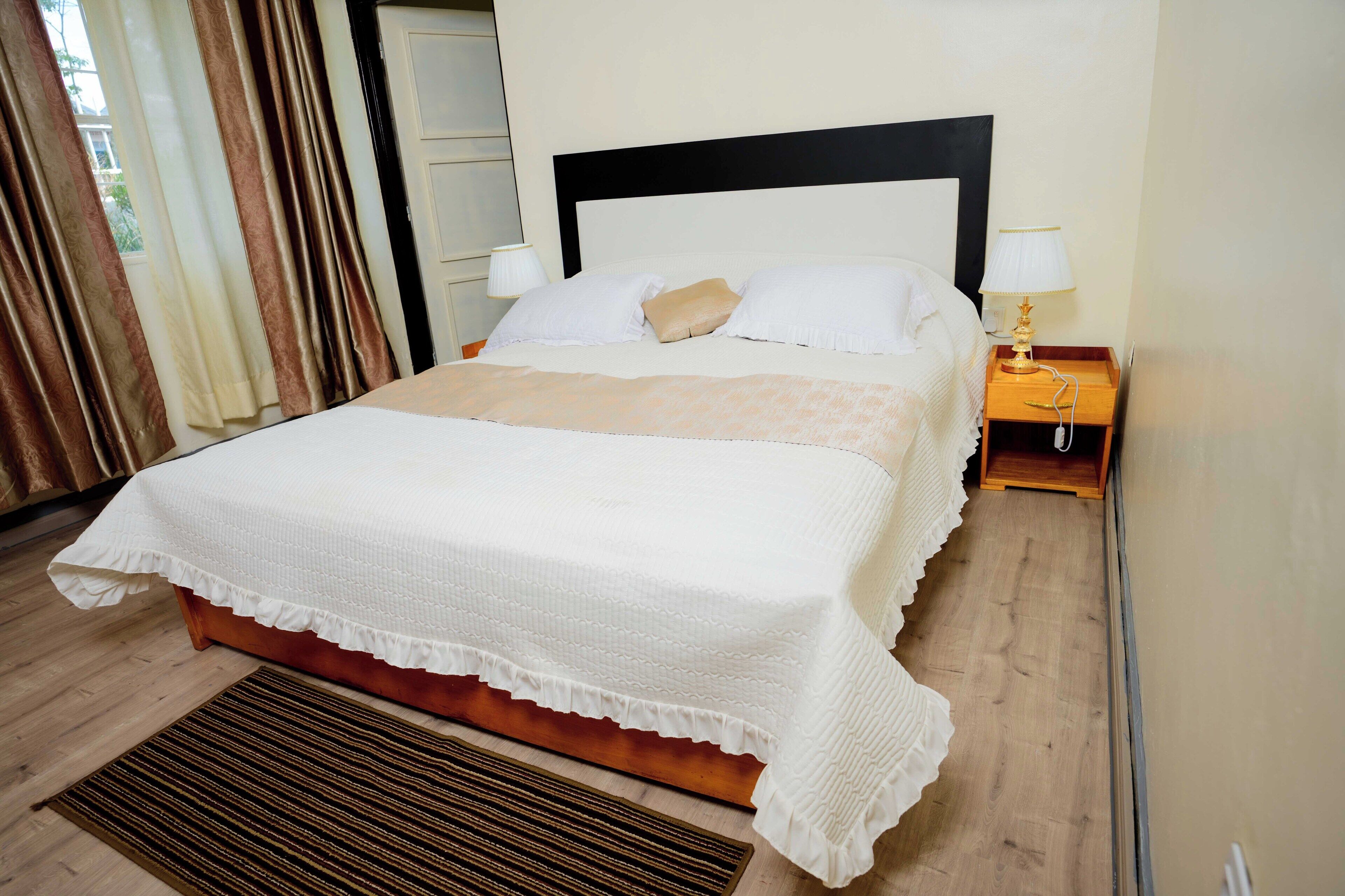 Una cama bien hecha con cubrecama y almohadas blancas, cabecera de madera, mesita de noche con lámpara y suelo de madera.