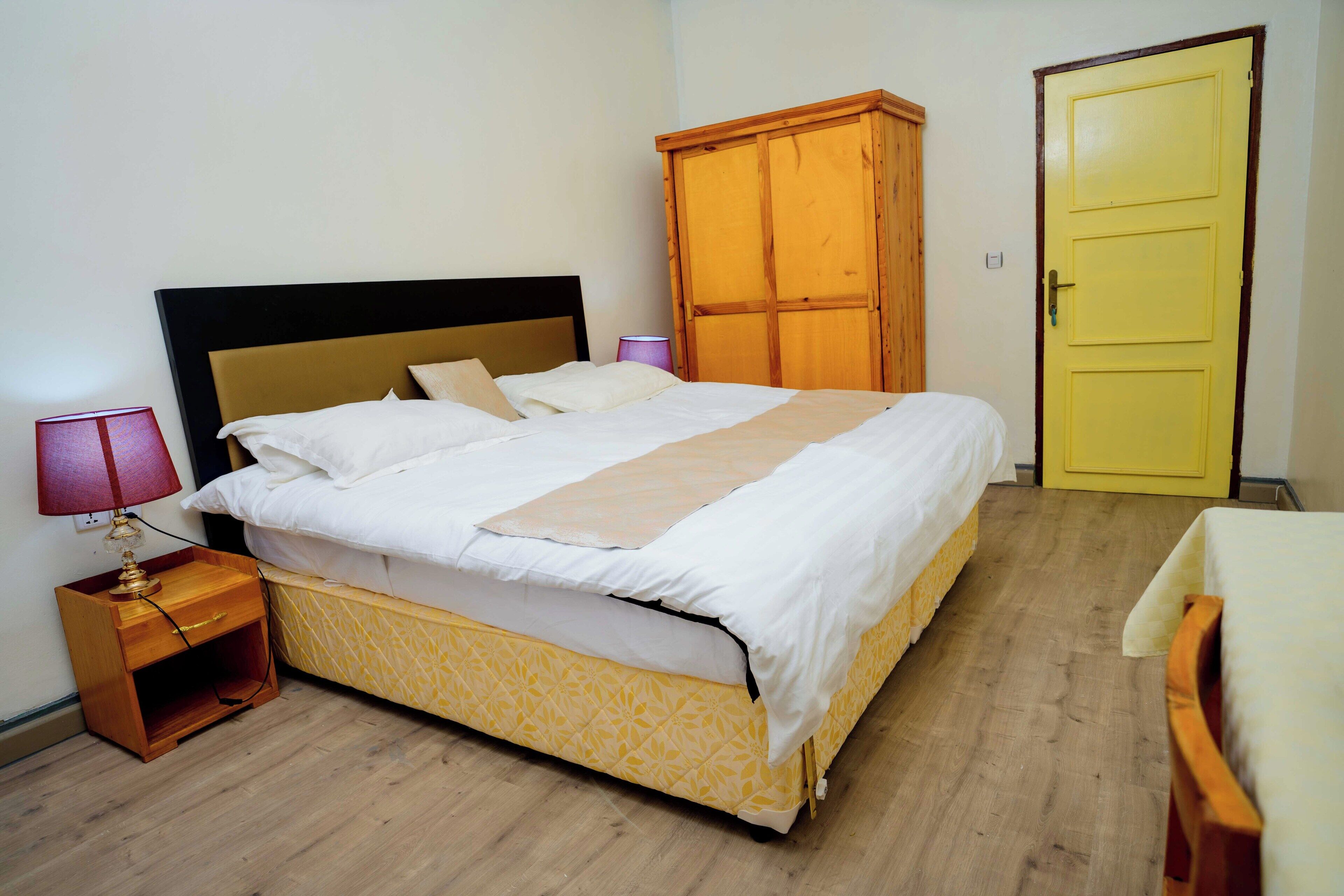 A neatly made bed with a patterned bedspread, a wooden nightstand with a lamp, and a yellow wardrobe in a room with wooden flooring.