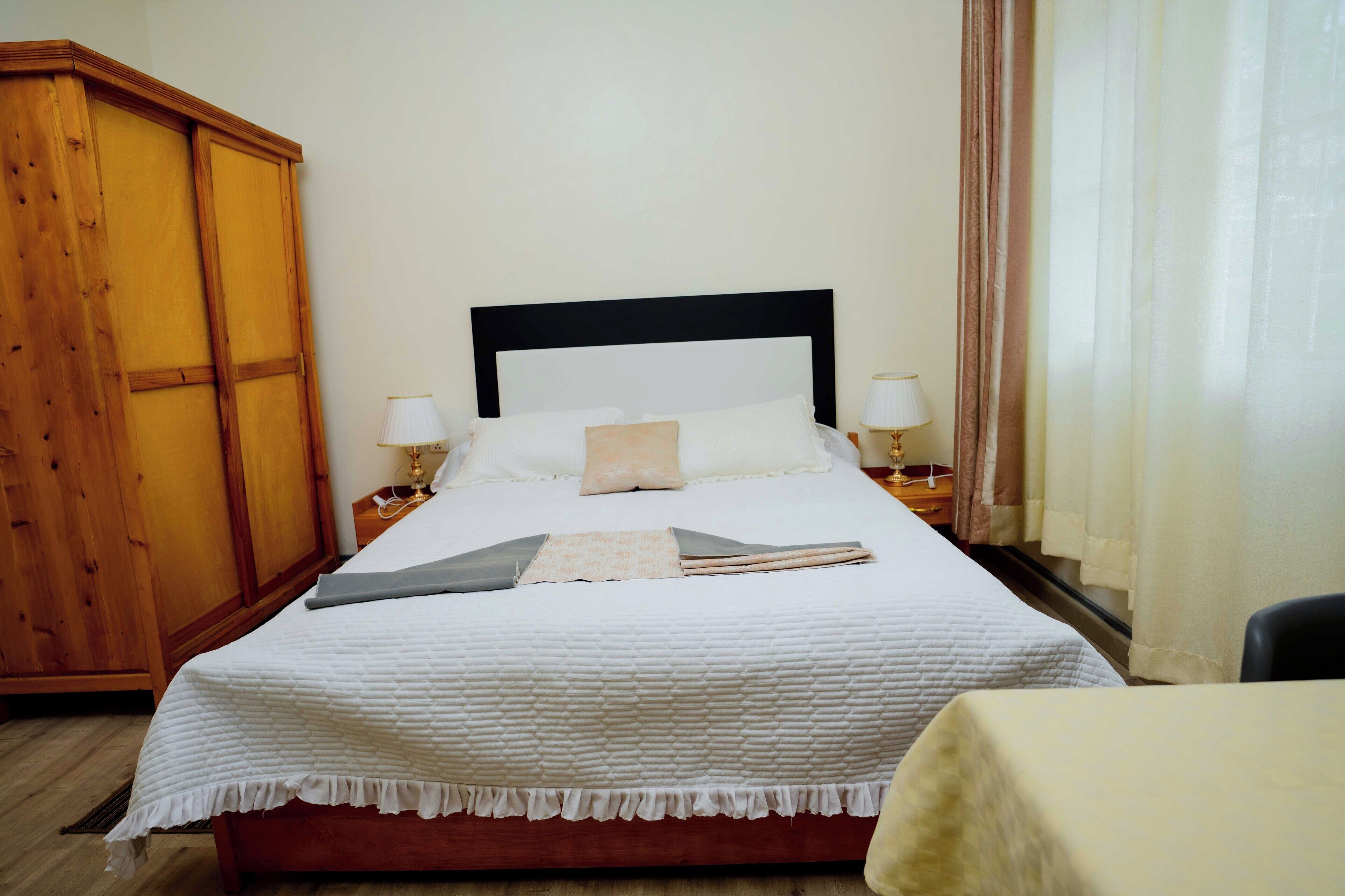 Een net opgemaakt bed met een witte dekbedovertrek, twee bedlampjes en een houten garderobe.
