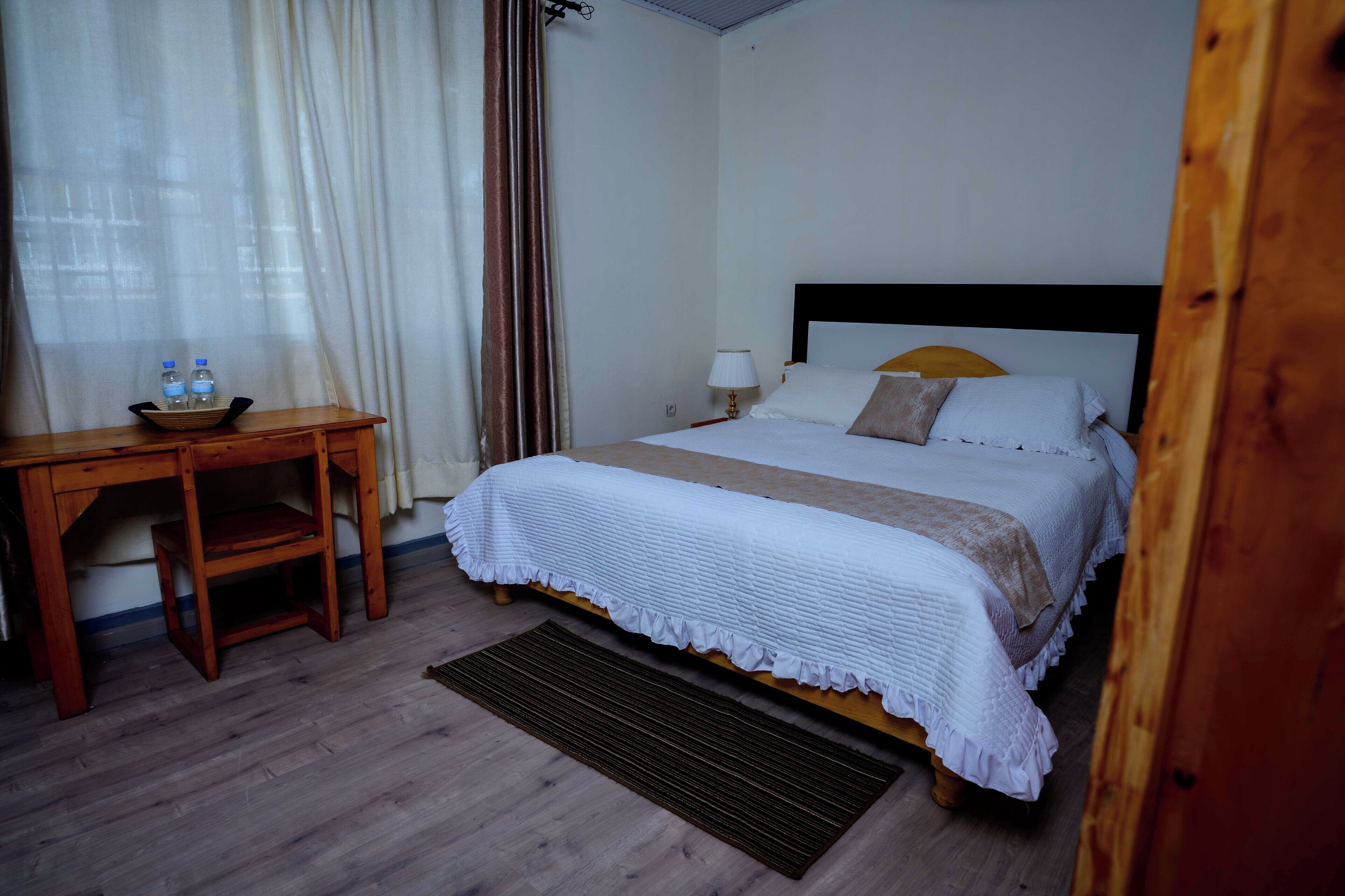 A hotel room with a bed, a desk with a chair, a small table, and a window with curtains.