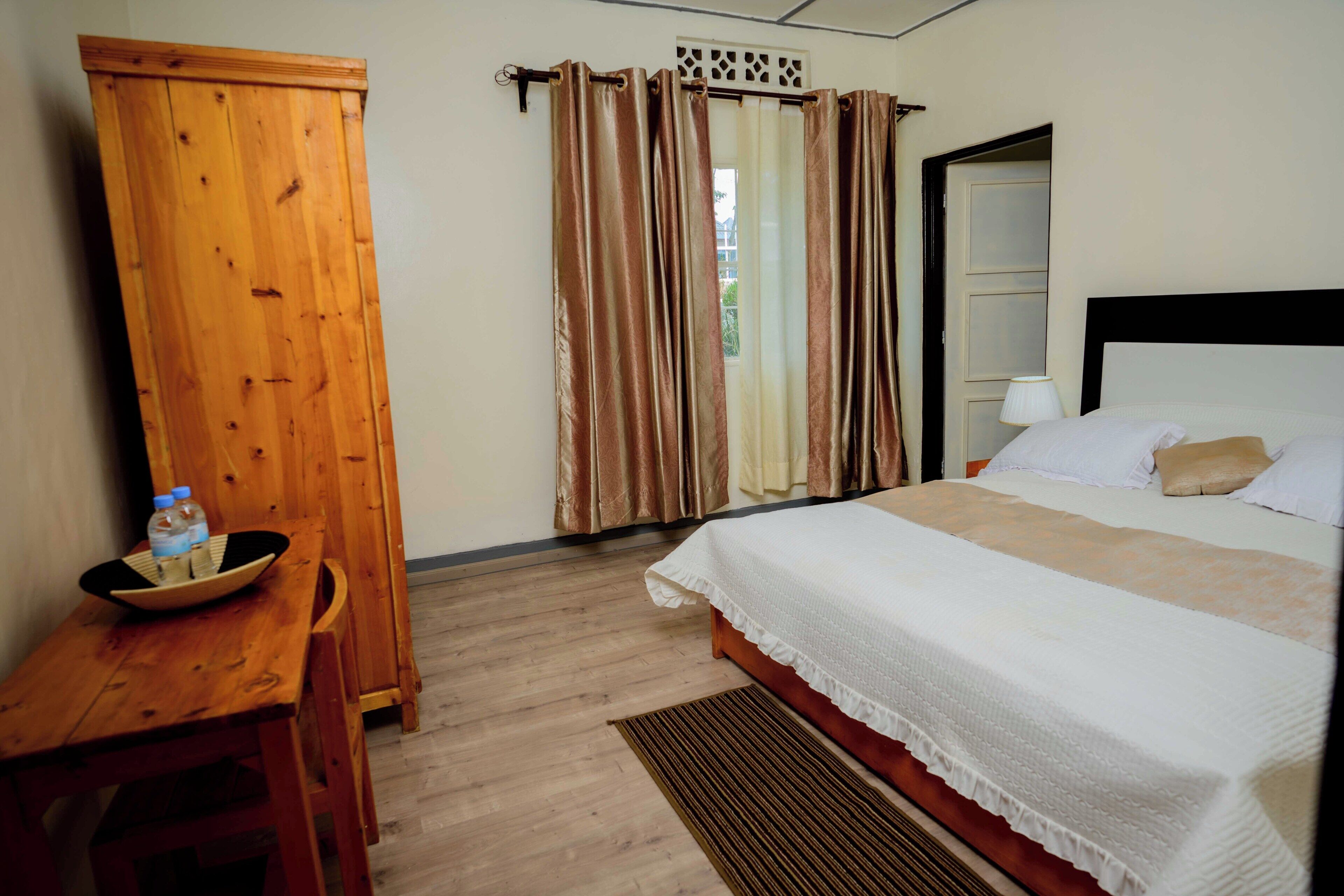 Una habitación de hotel con un armario de madera, una cama con ropa blanca, una mesita con un bol y botellas, y una ventana con cortinas.