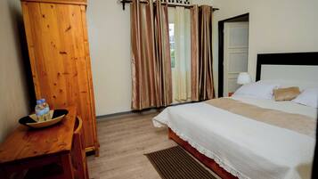 Una habitación de hotel con un armario de madera, una cama con ropa blanca, una mesita con un bol y botellas, y una ventana con cortinas.