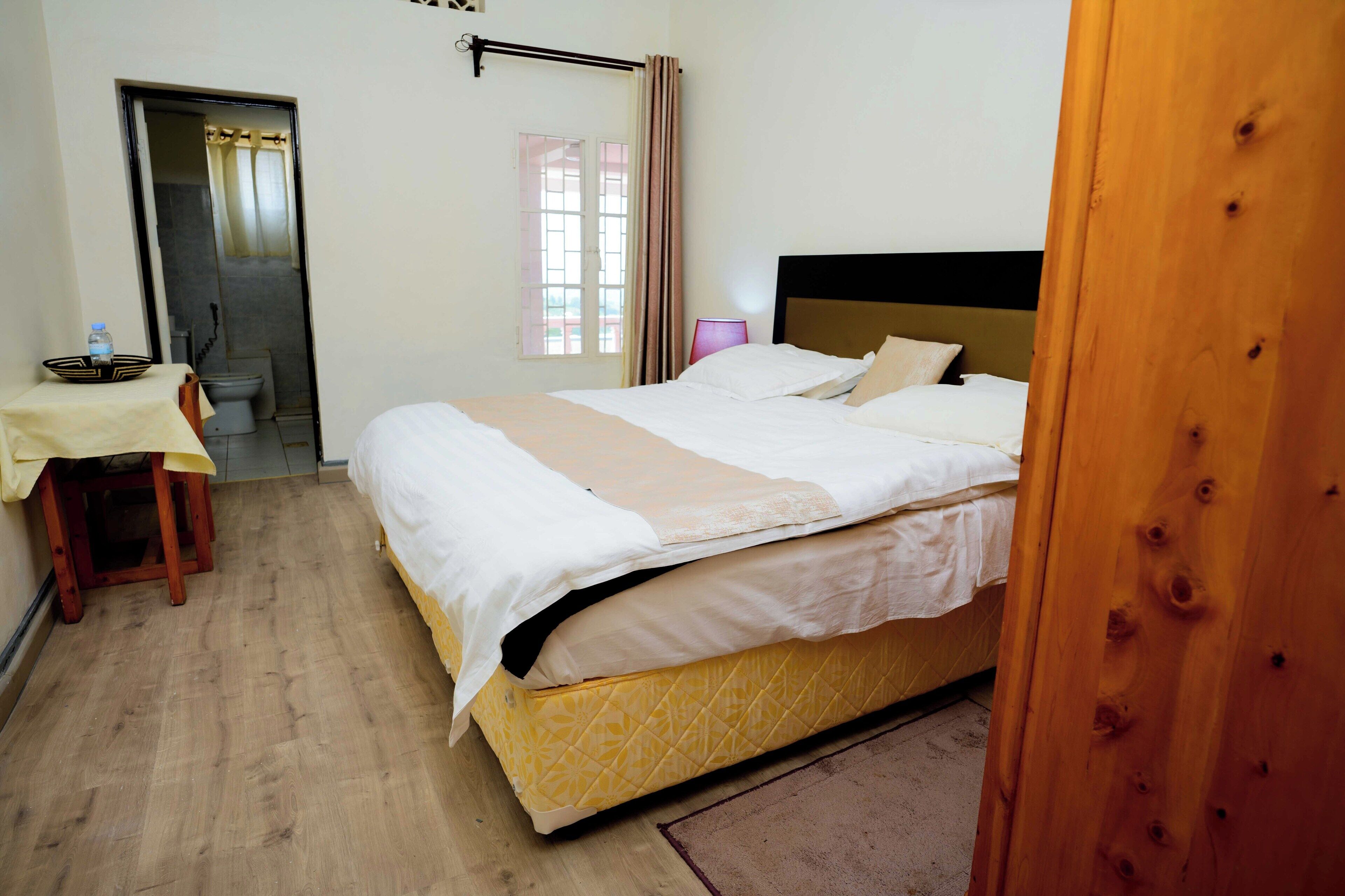 A hotel room with a bed, a small table with a water bottle, a chair, a window with curtains, and a door leading to a bathroom.