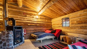 Ein Holzhaus-Interieur mit einem Kamin aus Stein, einem Bett mit roter und schwarzer Bettwäsche, einem kleinen Hocker und einem Fenster.