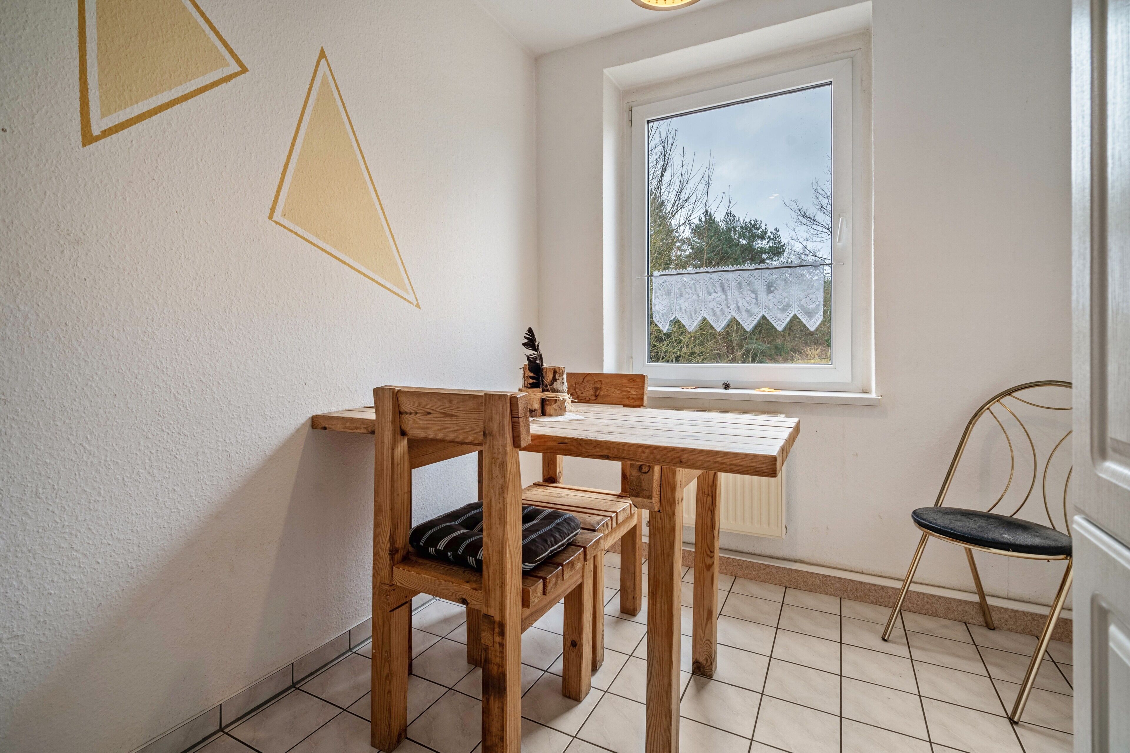 A room with a wooden table and chairs, a window with a view of trees, and a wall with geometric patterns.