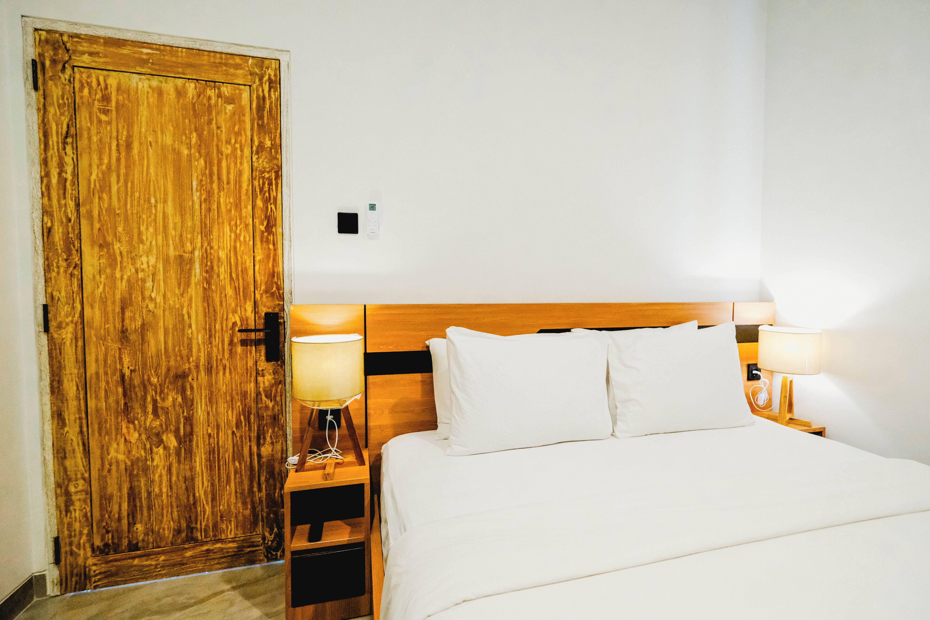 Habitación de hotel con puerta de madera, cama con ropa blanca, dos lámparas de noche y una mesita de noche.