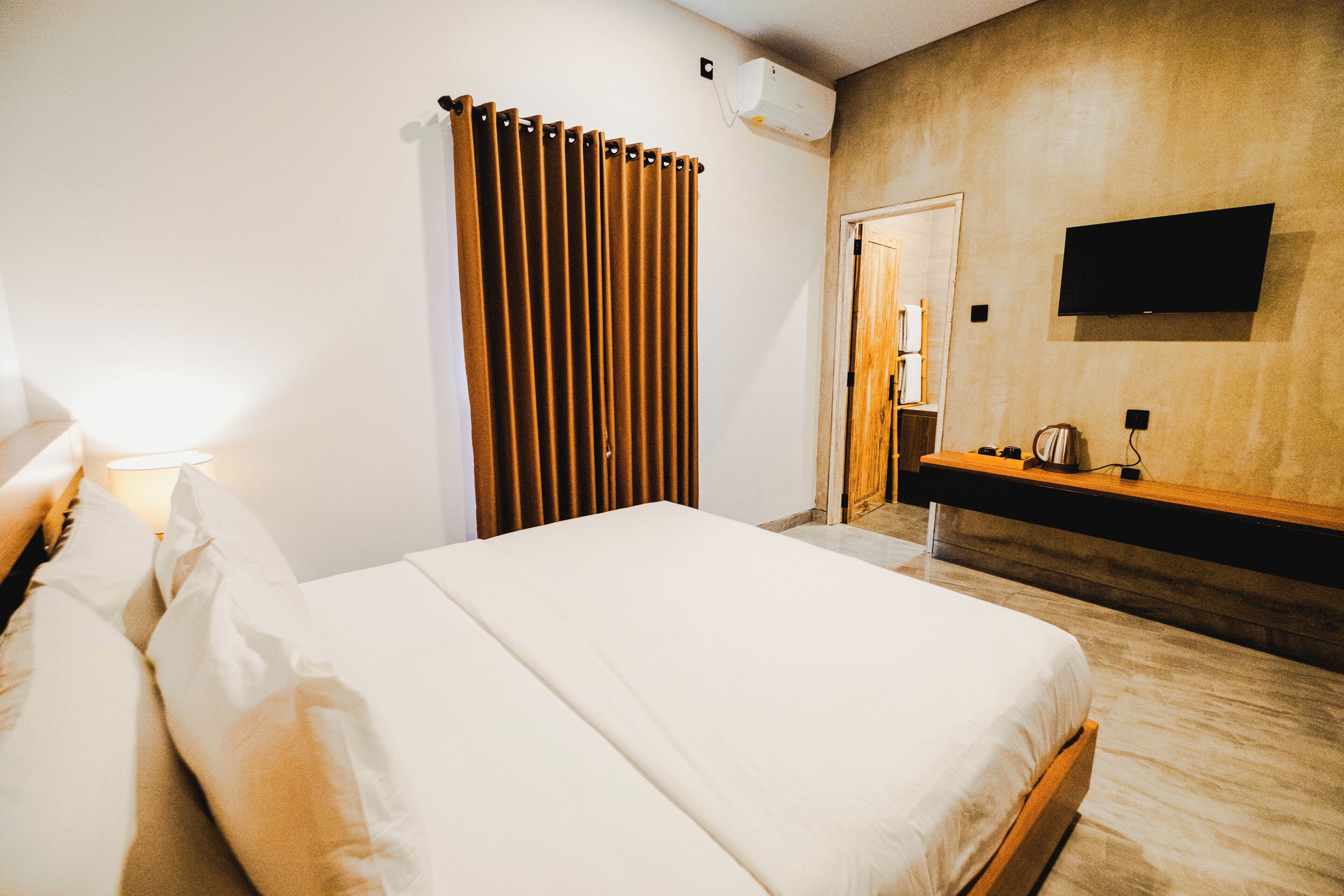 A hotel room with a bed, a bedside table, a TV mounted on the wall, and a window with curtains.