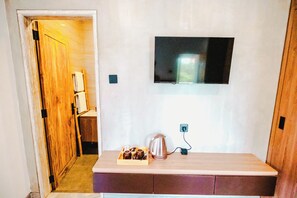 Una camera d'albergo con una porta in legno, una TV a schermo piatto fissata al muro, un tavolino con bollitore e tazze, e una presa elettrica.
