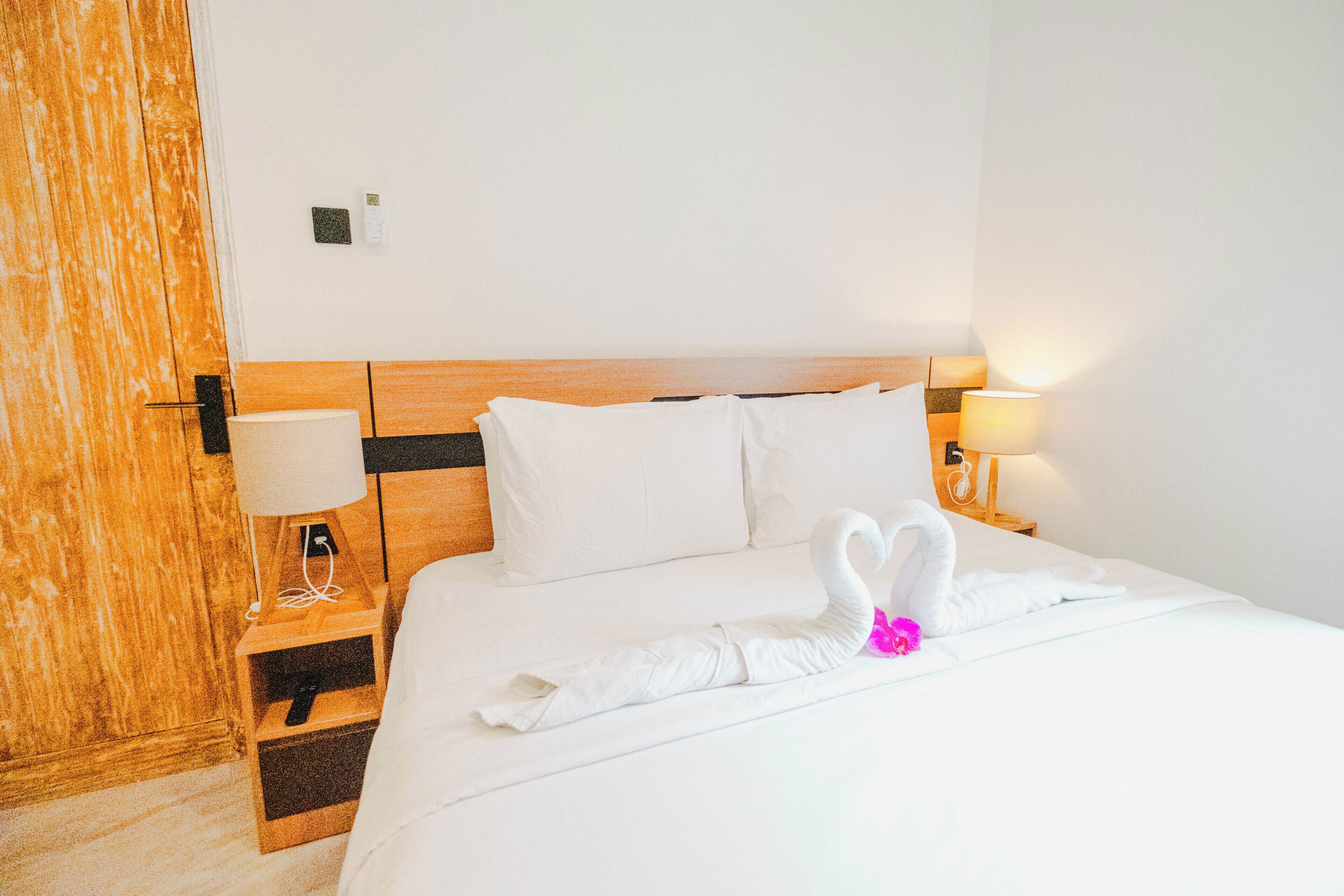 Habitación de hotel con cabecera de madera, ropa de cama blanca, lámpara en la mesita de noche y una toalla con forma de cisne sobre la cama.