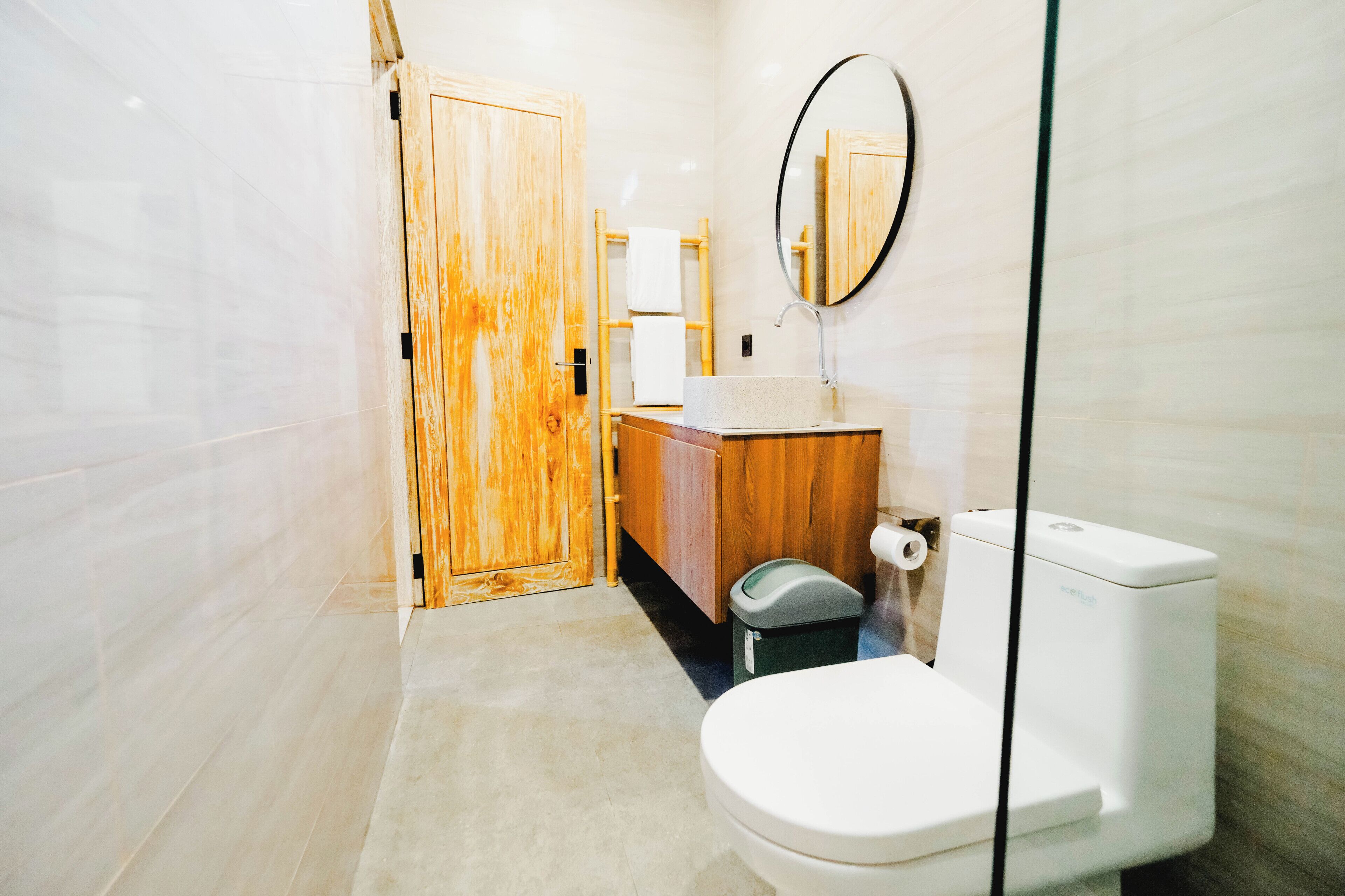 A bathroom with a wooden door, a round mirror, a toilet, and a sink with a towel rack.