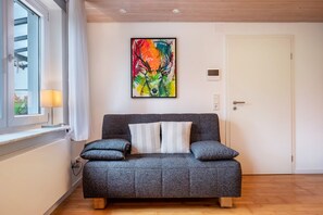 Un canapé gris avec deux coussins, un tableau abstrait aux couleurs vives accroché au mur, une porte blanche et une fenêtre avec une lampe.