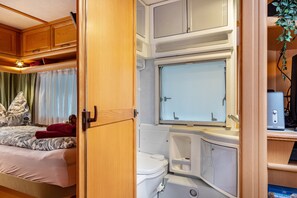 Interior compacto de autocaravana con cama, inodoro, lavamanos y ventana.