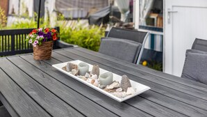 A wooden table with a white rectangular tray holding candles and decorative stones, a wicker basket with flowers, and a view of a garden and a building in the background. - Caravan 'La Pura Vida' with Private Terrace (Brandenburg an der Havel)