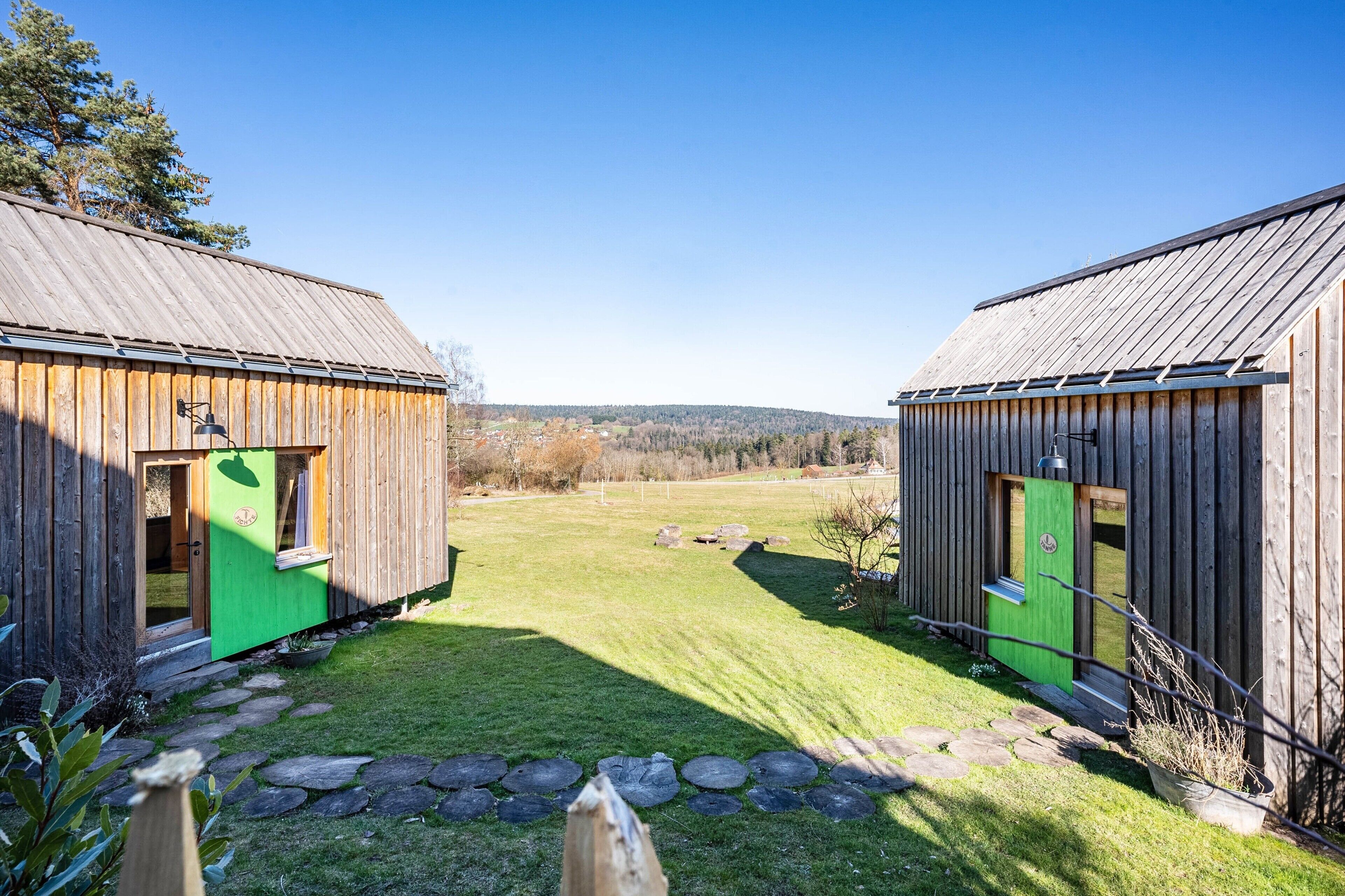 Two wooden cabins with green doors, a stone pathway, and a grassy yard.
