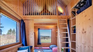 A wooden cabin interior with a loft bedroom, a window with a view, and a ladder to the loft.