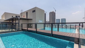 Una piscina en la azotea con barandilla de cristal, terraza de madera y vistas a edificios altos aledaños.