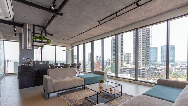 Una sala de estar moderna con un sofá, una mesa de centro y vistas a un paisaje urbano a través de amplios ventanales.
