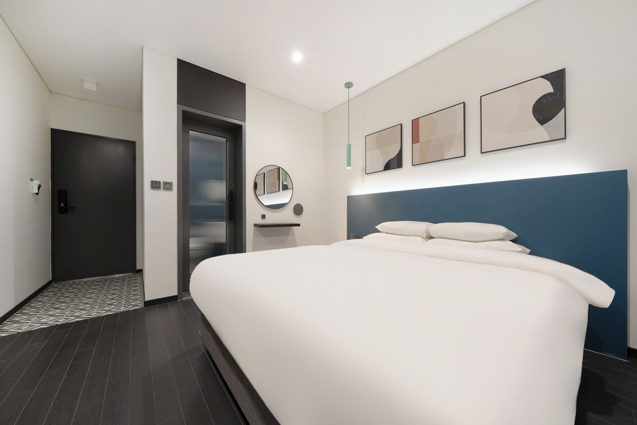 A modern hotel room with a large bed, a dark door, a small mirror, and abstract wall art.