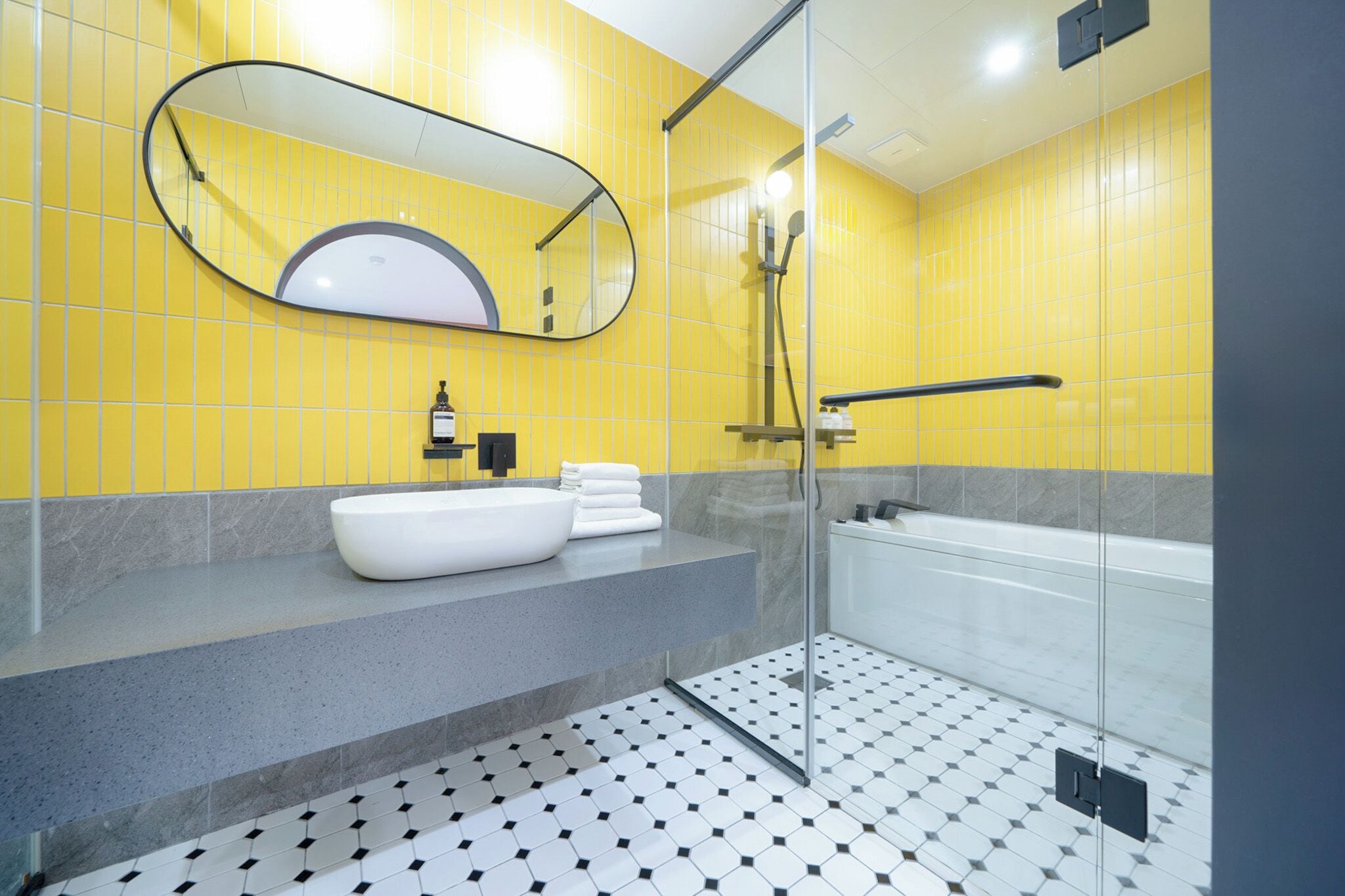A modern bathroom with a yellow tiled wall, a white sink, a large mirror, and a glass-enclosed shower area.
