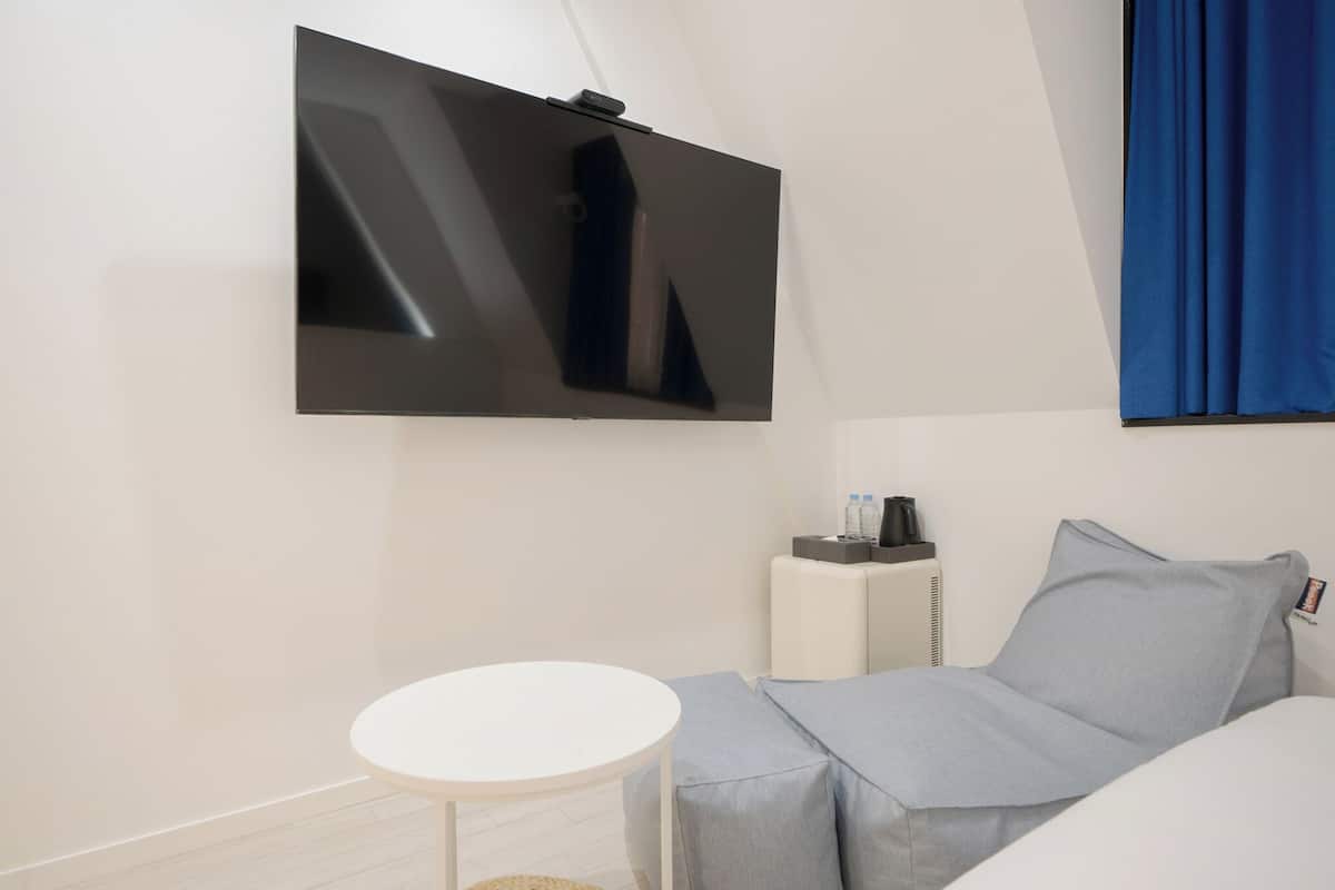 Ett modernt hotellrum med en platt-TV som är monterad på väggen, ett litet runt bord, en grå soffa och en blå gardin.