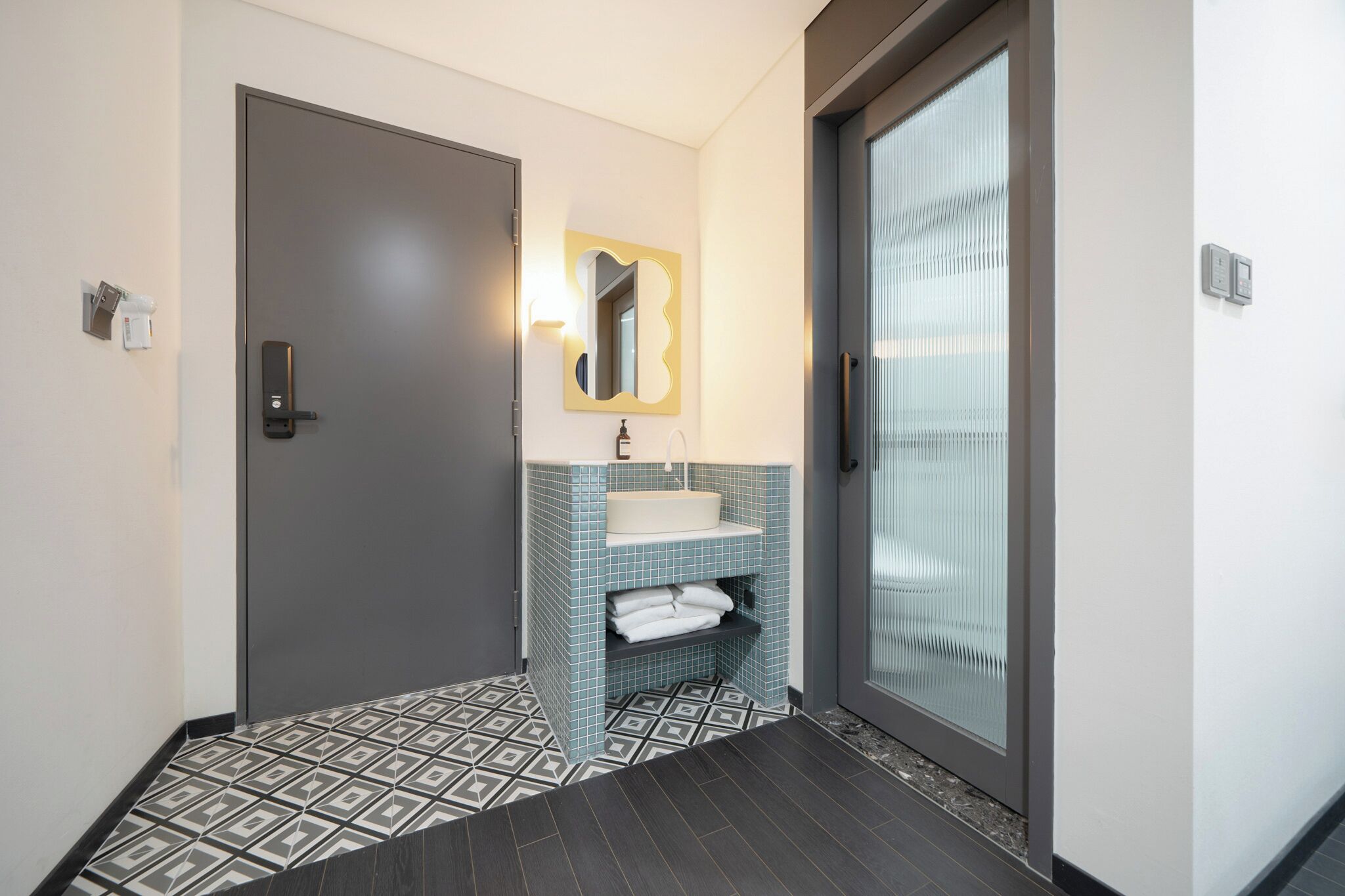 A modern bathroom with a grey door, a white sink with a blue tiled backsplash, and a mirror with a gold frame.