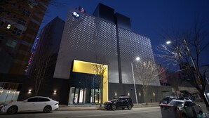 A modern building with a unique facade featuring a pattern of lights, a prominent entrance with glass doors, and a parking area with cars.