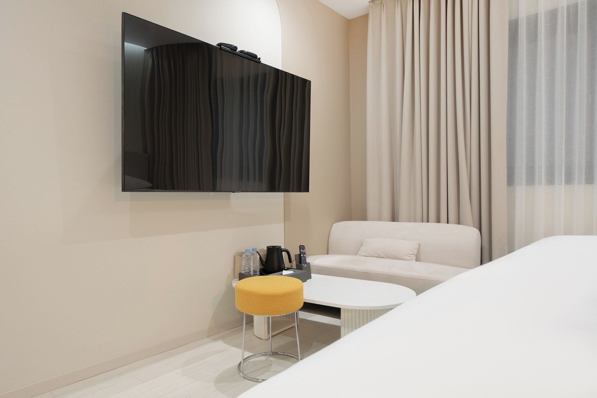 Habitación de hotel moderna con una televisión de pantalla plana montada en la pared, un sofá blanco, una pequeña mesa redonda con un taburete amarillo y un amplio ventanal con cortinas.