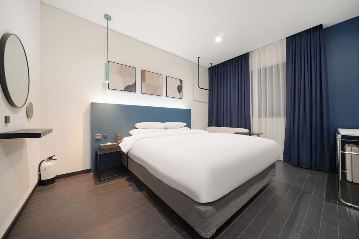 Ett modernt hotellrum med en stor säng, ett sängbord, en liten avlastningsyta, en spegel och ett fönster med gardiner.