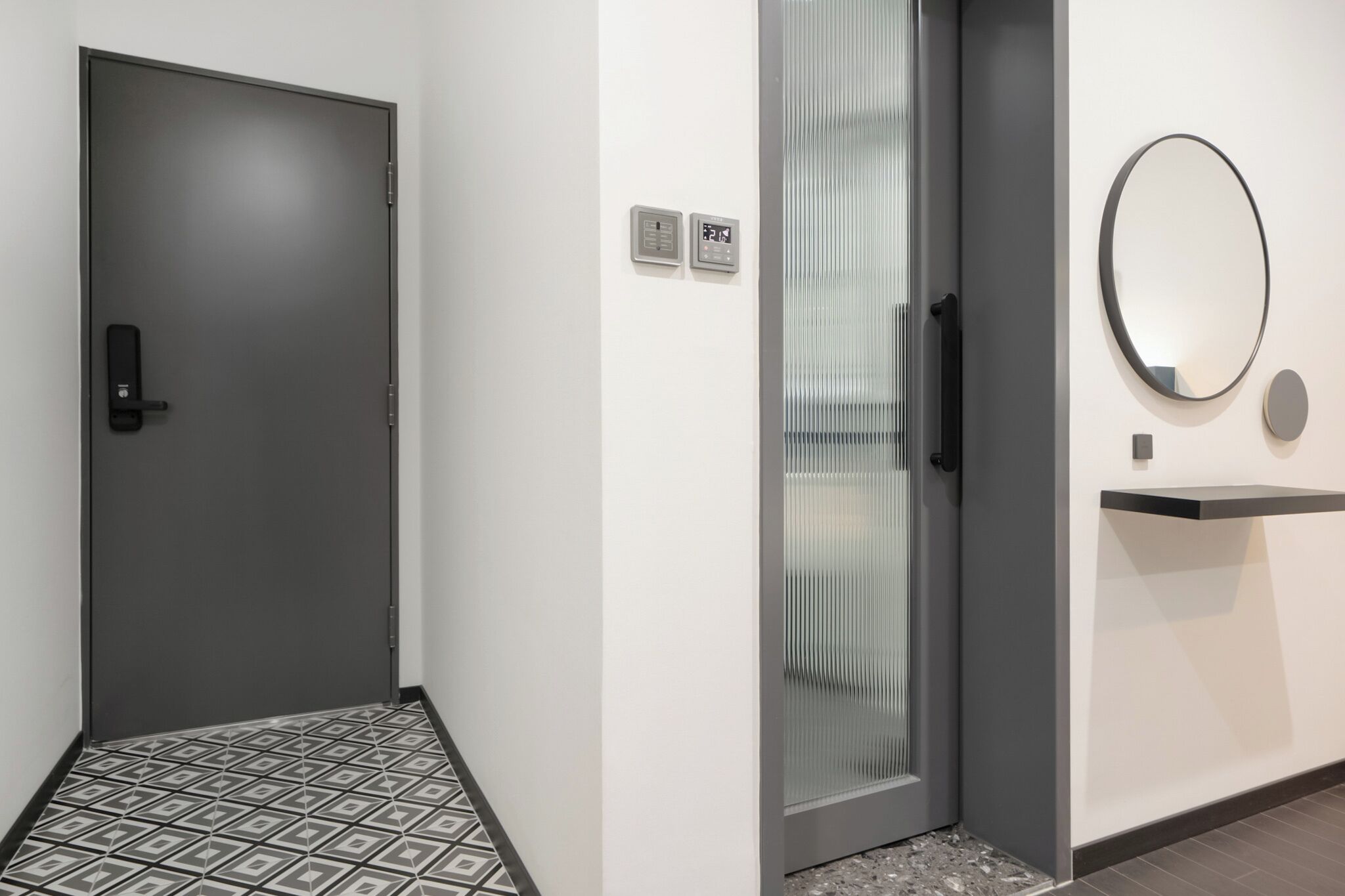 Un pasillo con una puerta oscura, una puerta de cristal con manija metálica y un espejo fijado a la pared.