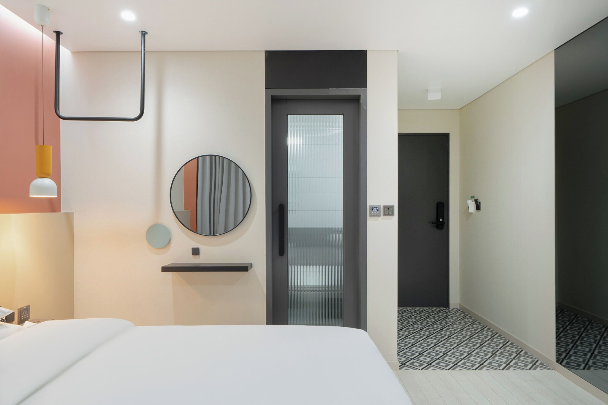 A modern hotel room with a bed, a round mirror, a small shelf, a door with a frosted glass panel, and a patterned floor.