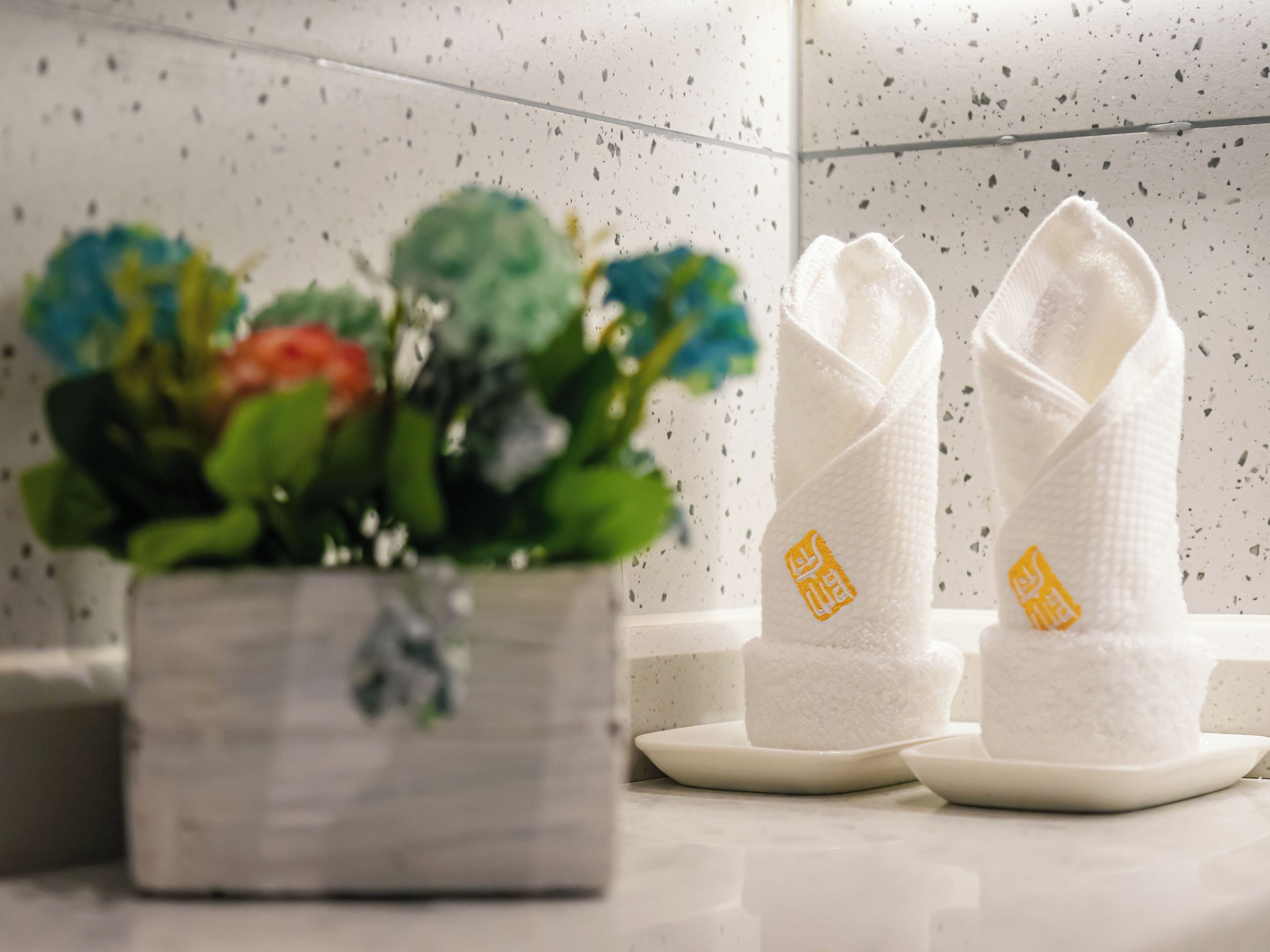 Uma casa de banho com uma pia branca, duas toalhas brancas com texto amarelo e uma planta em vaso com flores coloridas.