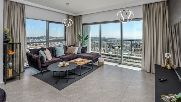 A modern living room with a large sofa, a coffee table, and a view of a cityscape through large windows.