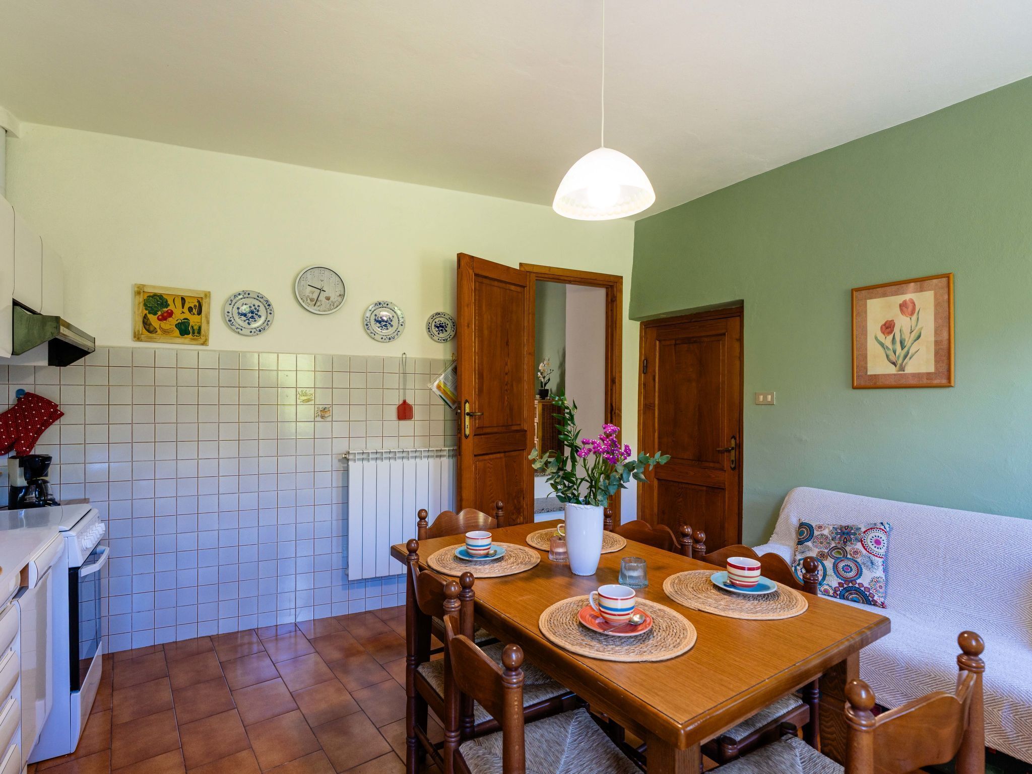 Un coin repas avec une table en bois dressée pour quatre personnes, entourée de chaises. Un radiateur blanc est fixé au mur, au-dessus du sol carrelé.