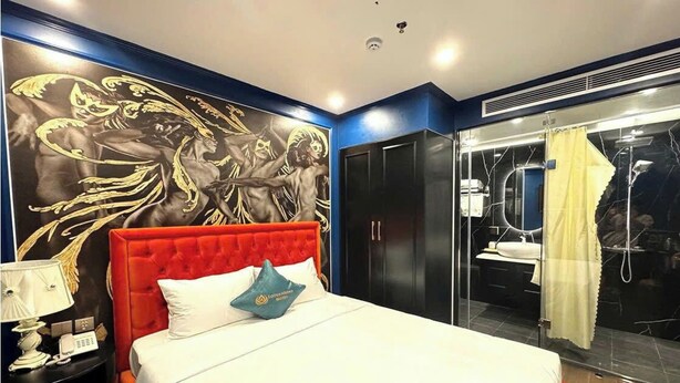 A hotel room with a red upholstered headboard, a large bed, a nightstand with a lamp, a wall mural, a bathroom with a glass shower, and a yellow curtain. - Lotus Aroma (Hanoi)