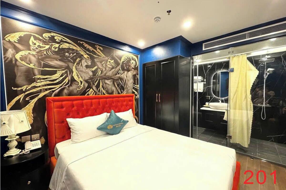 A hotel room with a red upholstered headboard, a large bed, a nightstand with a lamp, a wall mural, a bathroom with a glass shower, and a yellow curtain.