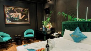 A hotel room with a bed, two teal chairs, a small table with a phone and a vase of flowers, and a painting on the wall. - Lotus Aroma (Hanoi)
