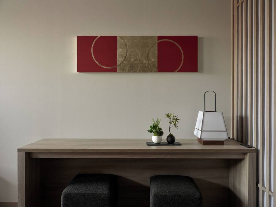 A modern desk with two black upholstered chairs, a small potted plant, and a lamp on a wooden table against a wall with a red and beige abstract painting.