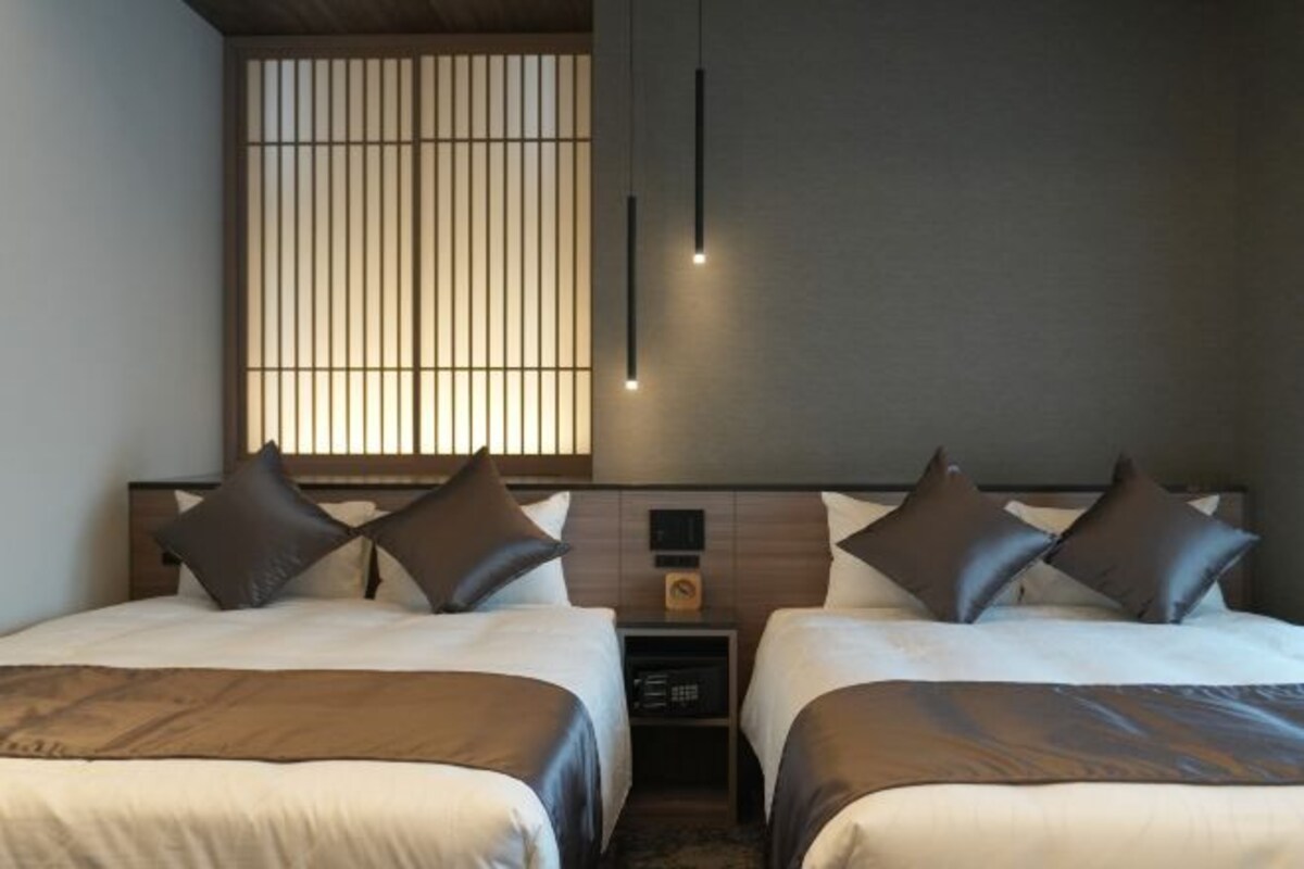 Habitación de hotel con dos camas, cabecero de madera, mesita de noche y ventana con persianas verticales.