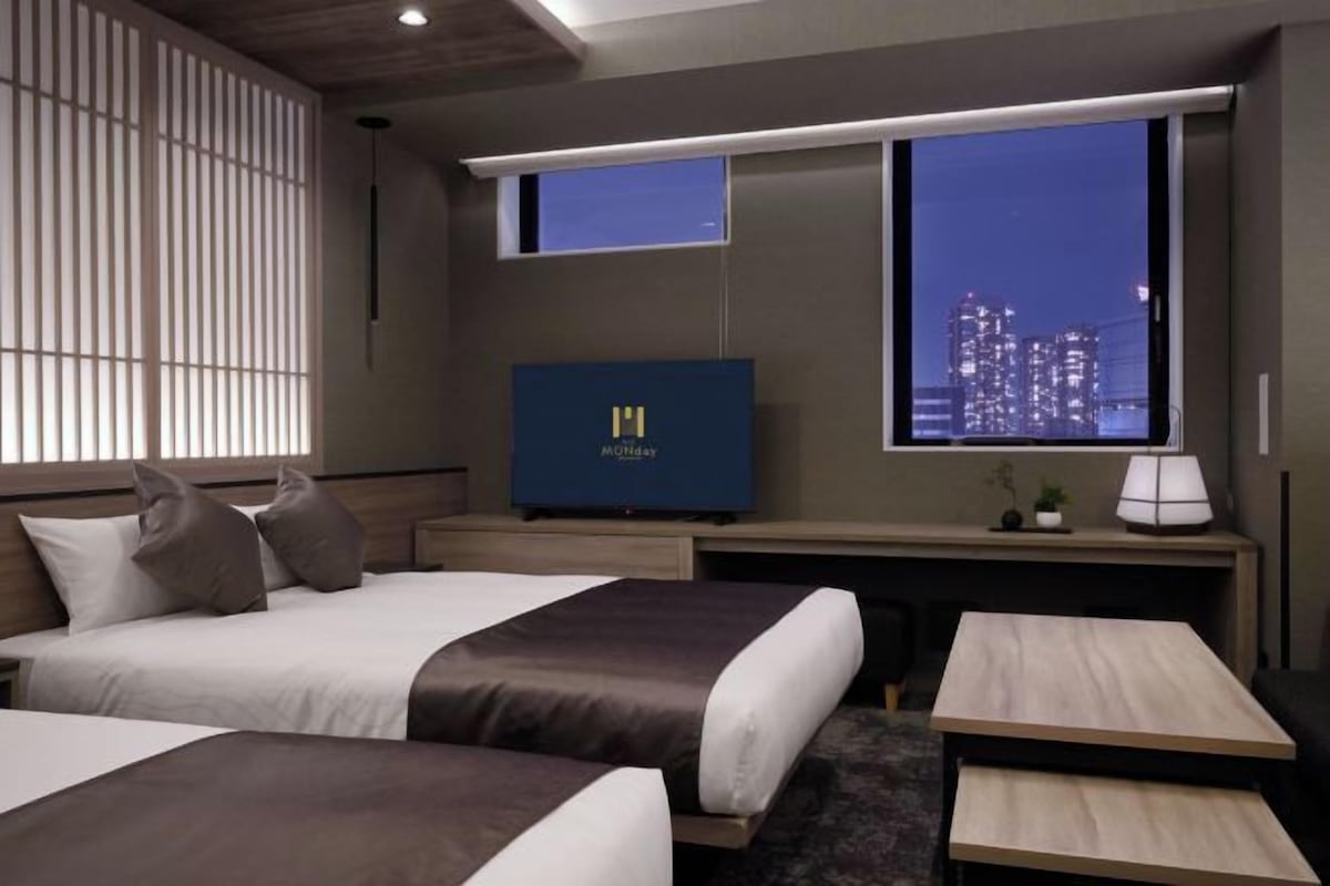 A hotel room with two beds, a desk, a TV, and a window with a city view.