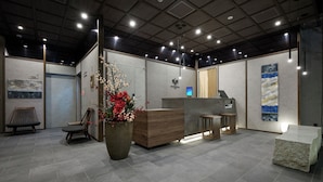 A reception area with a wooden reception desk, a large potted plant with red flowers, a seating area with chairs, and a stone sculpture. - GRAND MONday GINZA (Tokyo)